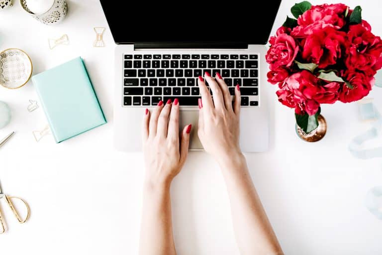 A person with red-painted nails types on a laptop at a white desk, surrounded by red flowers, a teal notebook, and gold paper clips—setting the scene for learning the basics of starting your own blog.