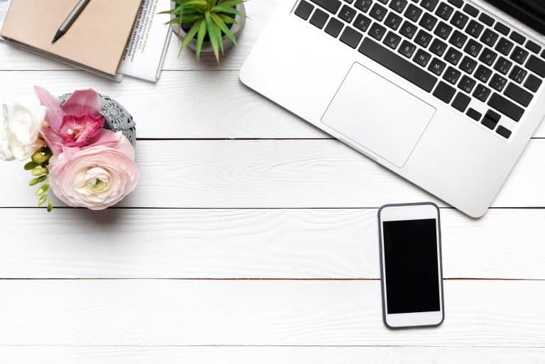 A laptop, smartphone, notebook with pen, potted plant, and a vase of flowers are arranged on a white wooden desk, leaving ample space in the center—perfect for working on your March Online Income Report.
